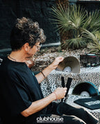 Person holding a cymbal and playing a drum set outdoors with plants in the background