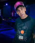 A man wearing a black t-shirt with a white line art graphic of a face and an orange circle, featuring the text 'CIRCO NERO ITALIA' at the bottom. He is also wearing a maroon trucker hat with 'CIRCO NERO' in white text. circo nero sorry im not a deejay