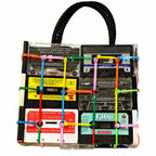 A novelty handbag designed to look like a clear plastic cassette-tape rack with a black braided top handle. The bag features multiple vintage cassette tapes in clear plastic, colorful plastic zip ties in green, pink, yellow, and blue, and labels from artists like Tabu and Daft Punk.