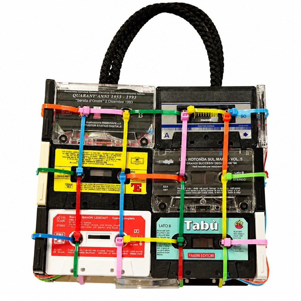 A novelty handbag designed to look like a clear plastic cassette-tape rack with a black braided top handle. The bag features multiple vintage cassette tapes in clear plastic, colorful plastic zip ties in green, pink, yellow, and blue, and labels from artists like Tabu and Daft Punk.
