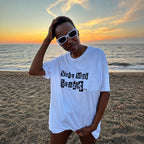 A person standing on a beach at sunset wearing an oversized white graphic T-shirt with black text reading 'None Mat Scap!'.