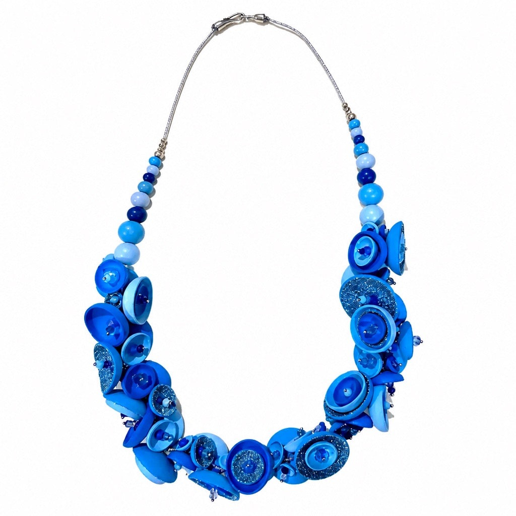 A vibrant blue statement necklace featuring a mix of flat round buttons, textured beads, and glass shell-like pieces in various shades of blue, suspended from a silver-tone chain on a white background.