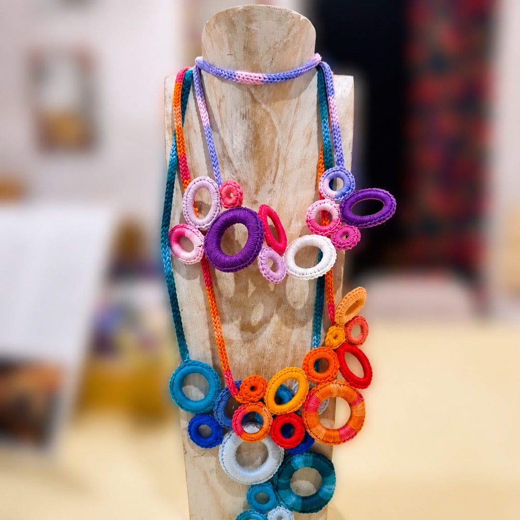 A colorful handmade beaded necklace with circular ring motifs displayed on a wooden stand. The necklace features multiple strands in vibrant shades of purple, blue, orange, pink, red, and white, set against a softly blurred indoor background. Silvia Tolomei best handmade jewelry in Tuscany. Tuscany jewelry. Tuscan jewelry. Populonia best store to visit. Things to do in populonia. 