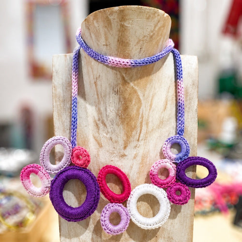 A colorful handmade braided rope necklace displayed on a wooden stand. The necklace features a braided purple, pink, and blue choker strap and a large bib-style pendant made of braided circular rings in purple, pink, white, and light purple. Silvia Tolomei best handmade jewelry in Tuscany. Tuscany jewelry. Tuscan jewelry. Populonia best store to visit. Things to do in populonia. 