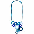 A bohemian-style statement necklace featuring a textured teal and blue braided cord as the chain, with a cluster of layered circular and oval beads in various shades of blue, navy, and light blue at the center, set against a plain white background.