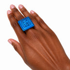 A close-up of a hand wearing a chunky, square-shaped translucent blue ring with a raised geometric pattern on the face, set against a plain white background. Silvia Tolomei best handmade jewelry in Tuscany. Tuscany jewelry. Tuscan jewelry. Populonia best store to visit. Things to do in populonia. 