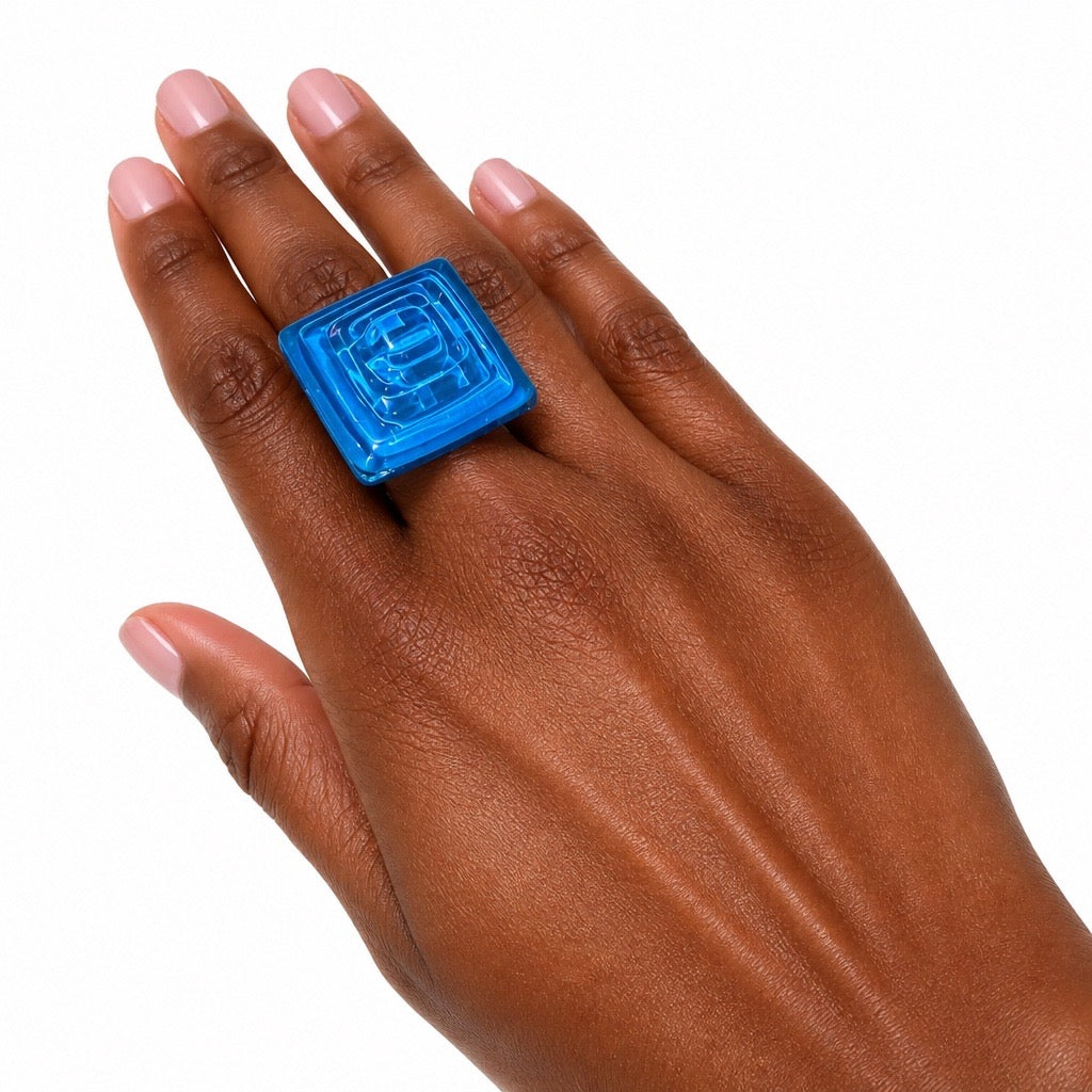 A close-up of a hand wearing a chunky, square-shaped translucent blue ring with a raised geometric pattern on the face, set against a plain white background. Silvia Tolomei best handmade jewelry in Tuscany. Tuscany jewelry. Tuscan jewelry. Populonia best store to visit. Things to do in populonia. 