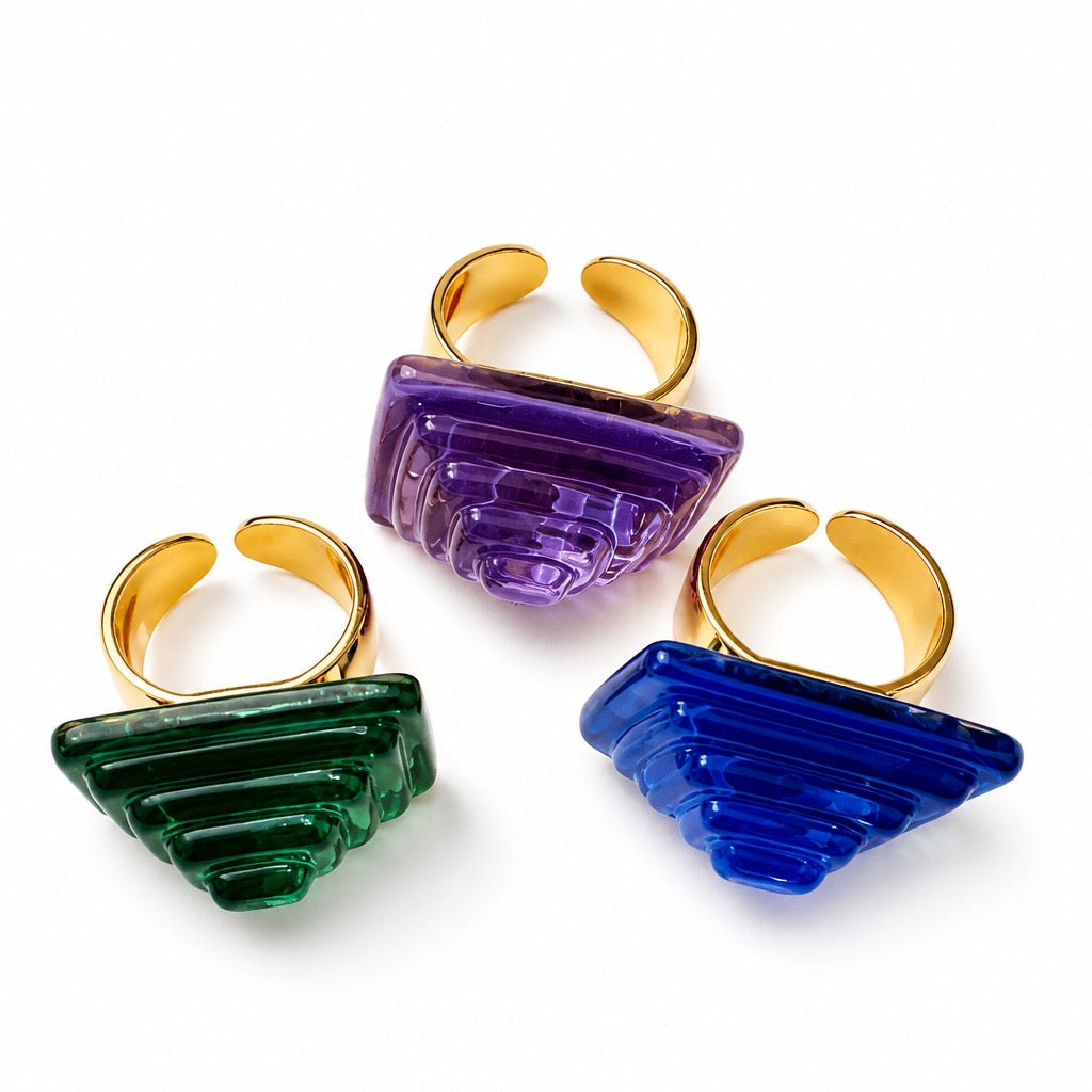 Three sculptural statement rings with gold-tone open bands and faceted translucent stone sections in green, purple, and blue, arranged on a white background. Silvia Tolomei best handmade jewelry in Tuscany. Tuscany jewelry.