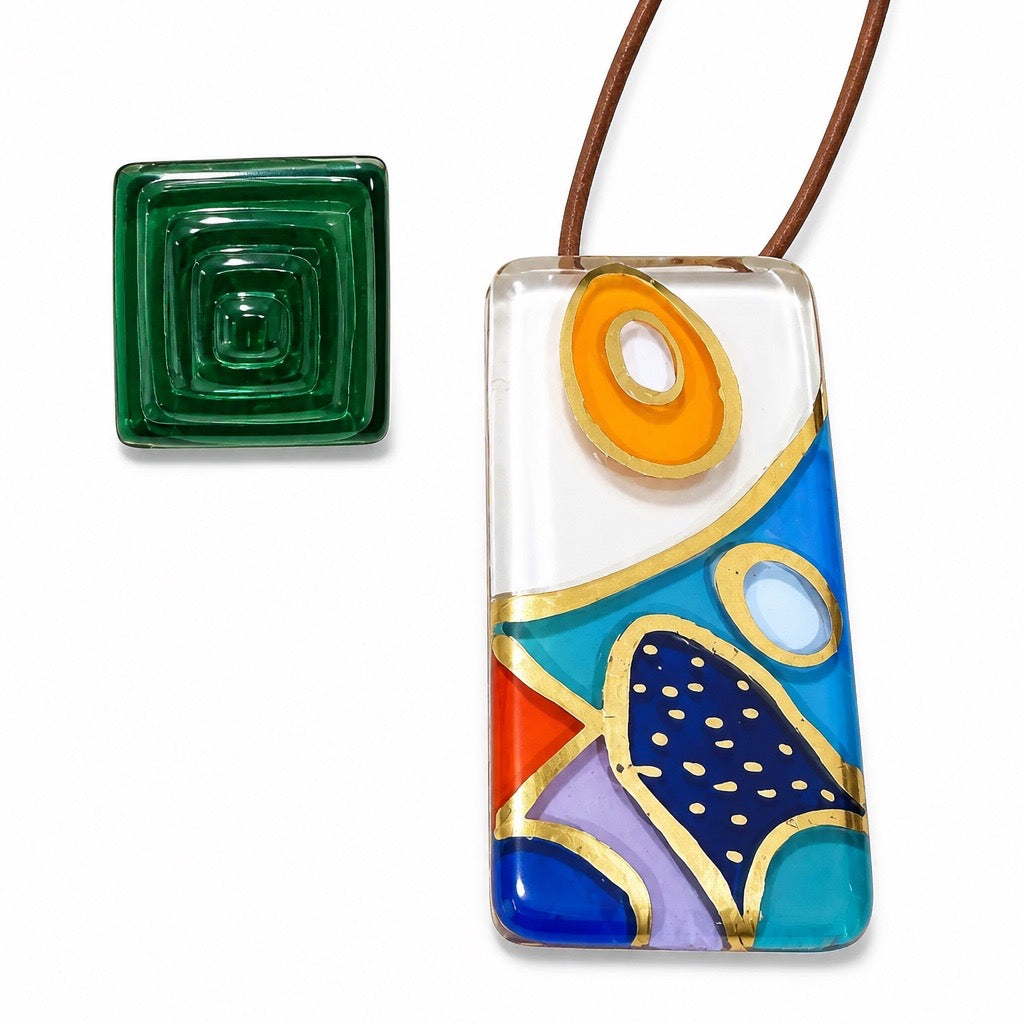 A rectangular glass pendant necklace with an abstract multicolor design on a clear glass base, featuring blue, orange, purple, red, and gold details, shown with a separate green square glass piece on a white background. Silvia Tolomei best handmade jewelry in Tuscany. Tuscany jewelry. Tuscan jewelry. Populonia best store to visit. Things to do in populonia. 