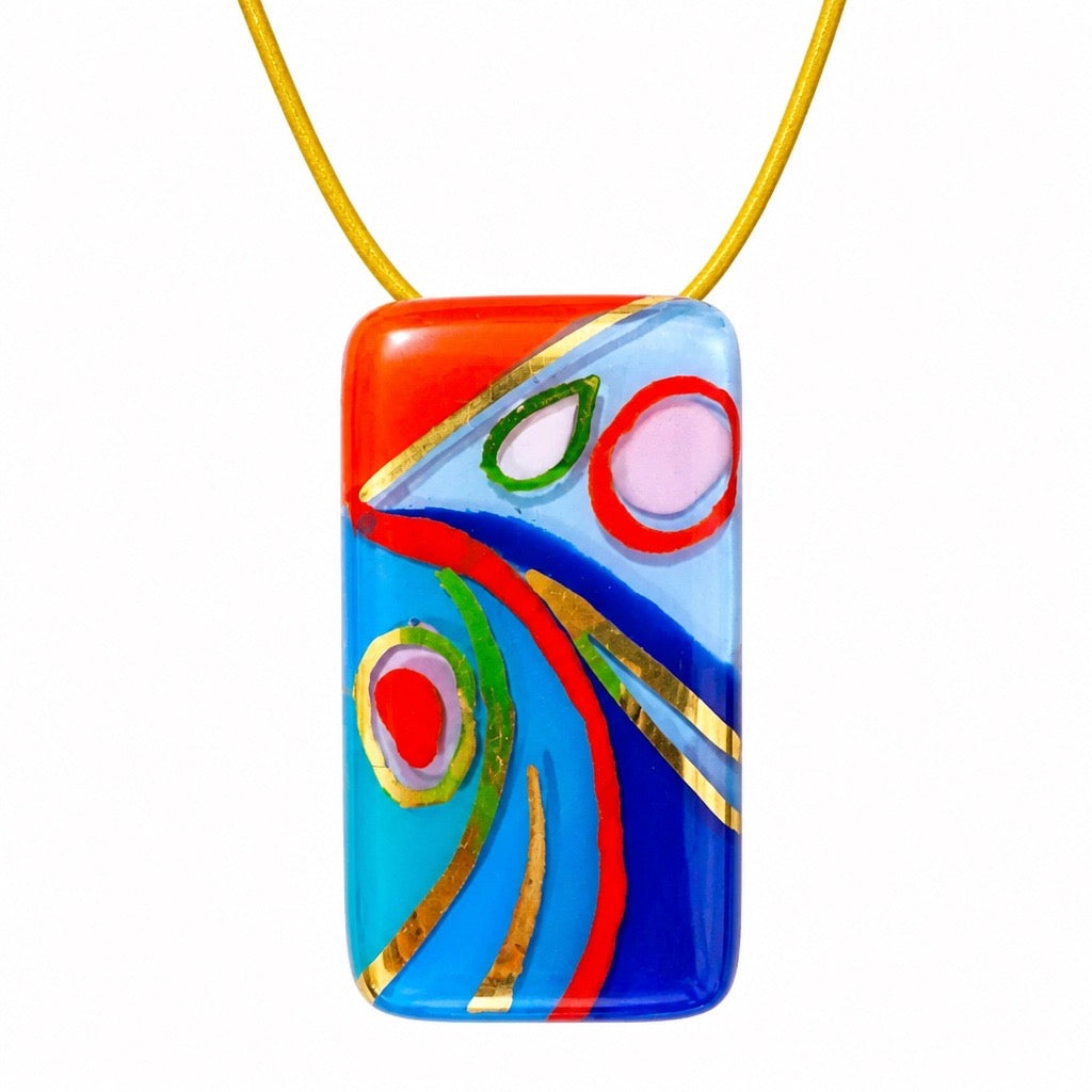 A colorful rectangular glass pendant necklace with abstract painted patterns in red, blue, light blue, pink, green, and gold, suspended from a yellow cord on a white background. Silvia Tolomei best handmade jewelry in Tuscany. Tuscany jewelry. Tuscan jewelry. Populonia best store to visit. Things to do in populonia. 