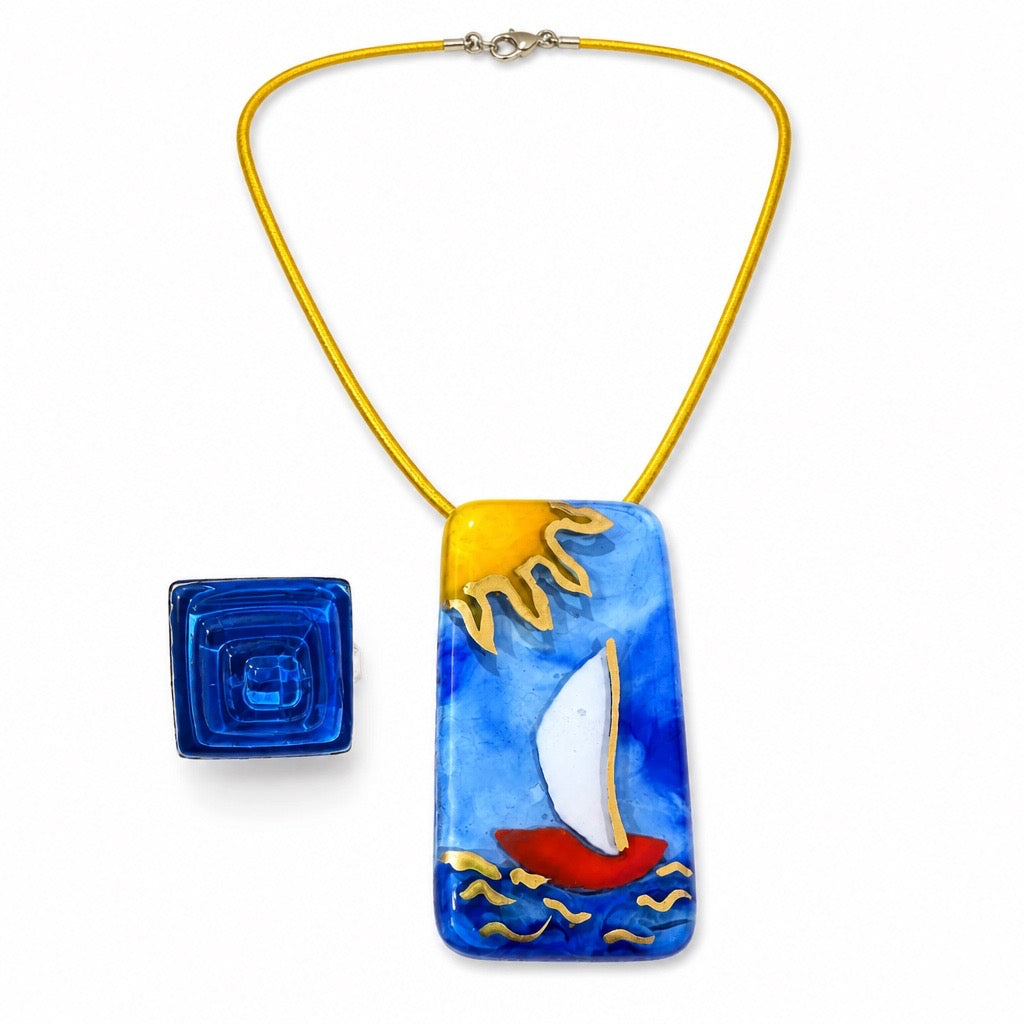 A handmade glass pendant necklace featuring a colorful sailboat design on a blue water background, suspended from a bright yellow cord. A small blue square glass charm is shown next to the pendant on a clean white background. Silvia Tolomei best handmade jewelry in Tuscany. Tuscany jewelry. Tuscan jewelry. Populonia best store to visit. Things to do in populonia. 