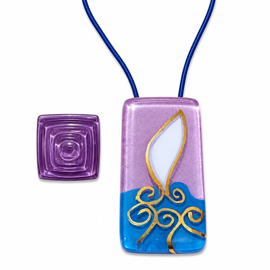 A rectangular fused glass pendant necklace with a blue cord, featuring a lavender top half, a bright blue bottom half, a white flame motif outlined in gold, and gold scrollwork. A small square purple glass bead is shown to the left on a white background. Silvia Tolomei best handmade jewelry in Tuscany. Tuscany jewelry. Tuscan jewelry. Populonia best store to visit. Things to do in populonia. 