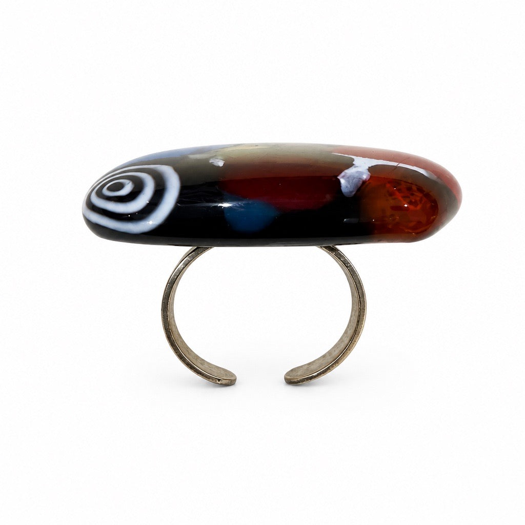 A handmade lampwork ring featuring a stylized fish design with a black and white spiral pattern on the left, a red and brown gradient body, and a blue accent, mounted on a silver-tone metal band. Silvia Tolomei best handmade jewelry in Tuscany. Tuscany jewelry. Tuscan jewelry. Populonia best store to visit. Things to do in populonia. 