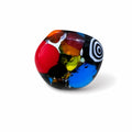 A round, hand-blown glass bead featuring a multicolor design with a large red section, a blue section, a black base with white spiral pattern, and orange and yellow flecks, shown on a white background. Silvia Tolomei best handmade jewelry in Tuscany. Tuscany jewelry. Tuscan jewelry. Populonia best store to visit. Things to do in populonia. 