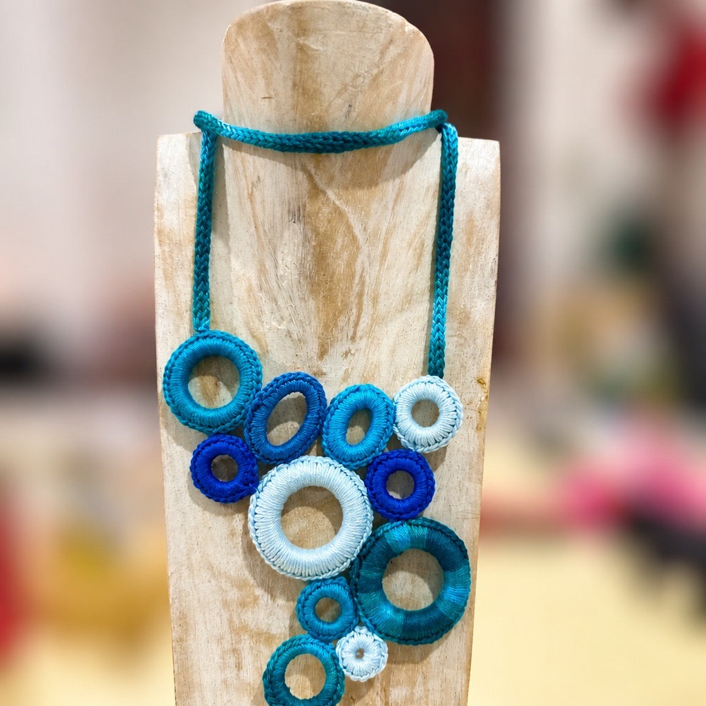A handmade bohemian-style necklace made of knotted turquoise, blue, and white rope rings, displayed on a light-colored wooden stand.