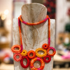 A chunky woven statement necklace with circular and oval links in vibrant orange, red, pink, and yellow, displayed on a light-colored wooden mannequin bust. Silvia Tolomei best handmade jewelry in Tuscany. Tuscany jewelry. Tuscan jewelry. Populonia best store to visit. Things to do in populonia. 