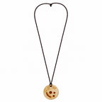 A circular wooden pendant necklace with a carved smiley face design, suspended on a dark brown cord against a plain white background. Silvia Tolomei best handmade jewelry in Tuscany. Tuscany jewelry. Tuscan jewelry. Populonia best store to visit. Things to do in populonia. 