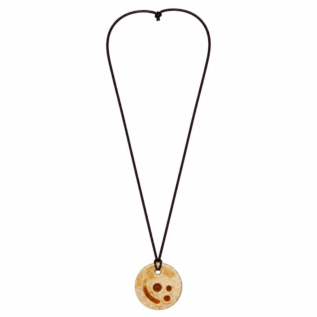 A circular wooden pendant necklace with a carved smiley face design, suspended on a dark brown cord against a plain white background. Silvia Tolomei best handmade jewelry in Tuscany. Tuscany jewelry. Tuscan jewelry. Populonia best store to visit. Things to do in populonia. 