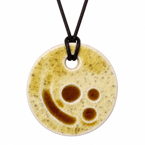 A round ceramic pendant necklace featuring a crackled glaze in cream and light brown with a stylized smiley face design, suspended on a black cord against a white background. Silvia Tolomei best handmade jewelry in Tuscany. Tuscany jewelry. Tuscan jewelry. Populonia best store to visit. Things to do in populonia. 