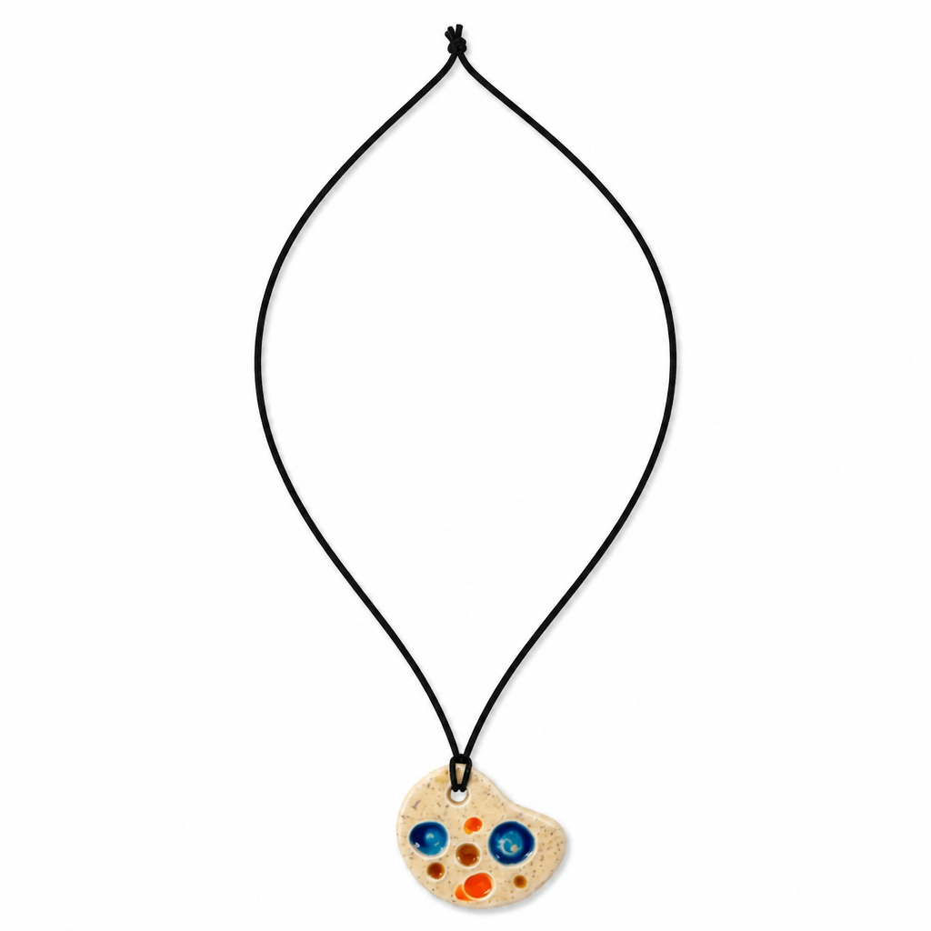 A black cord necklace featuring a wooden paint palette pendant with colorful painted dots in blue, orange, and red on a white background. Silvia Tolomei best handmade jewelry in Tuscany. Tuscany jewelry. Tuscan jewelry. Populonia best store to visit. Things to do in populonia. 