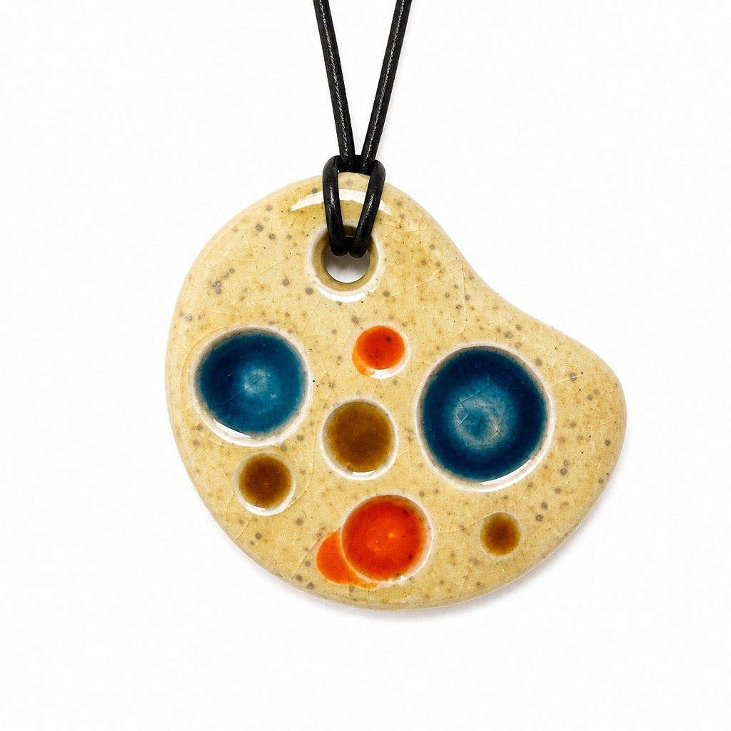 An irregularly shaped ceramic pendant necklace with a speckled beige glaze and colorful abstract circular patterns in blue, orange, and brown, hanging from a black cord on a white background. Silvia Tolomei best handmade jewelry in Tuscany. Tuscany jewelry. Tuscan jewelry. Populonia best store to visit. Things to do in populonia. 