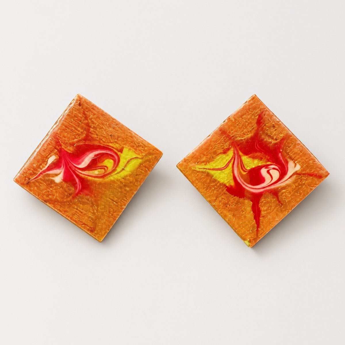 A pair of square stud earrings with a textured orange and yellow base and a red and white swirl design in the center, displayed on a clean white background. Silvia Tolomei best handmade jewelry in Tuscany. Tuscany jewelry. Tuscan jewelry. Populonia best store to visit. Things to do in populonia. 