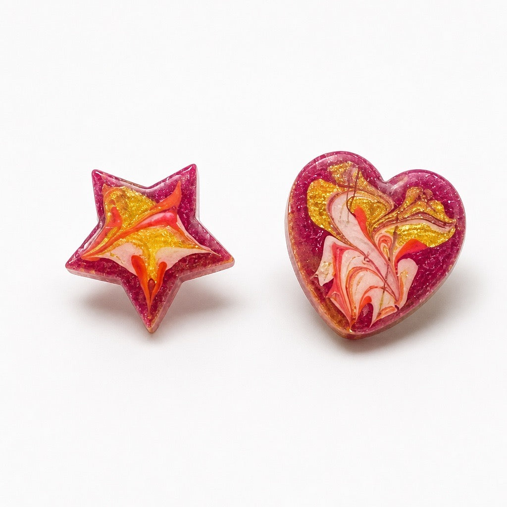 A pair of handmade resin stud earrings on a white background, featuring one star-shaped earring and one heart-shaped earring, both with a marbled swirl pattern of pink, yellow, orange, and white. Silvia Tolomei best handmade jewelry in Tuscany. Tuscany jewelry. Tuscan jewelry. Populonia best store to visit. Things to do in populonia. 