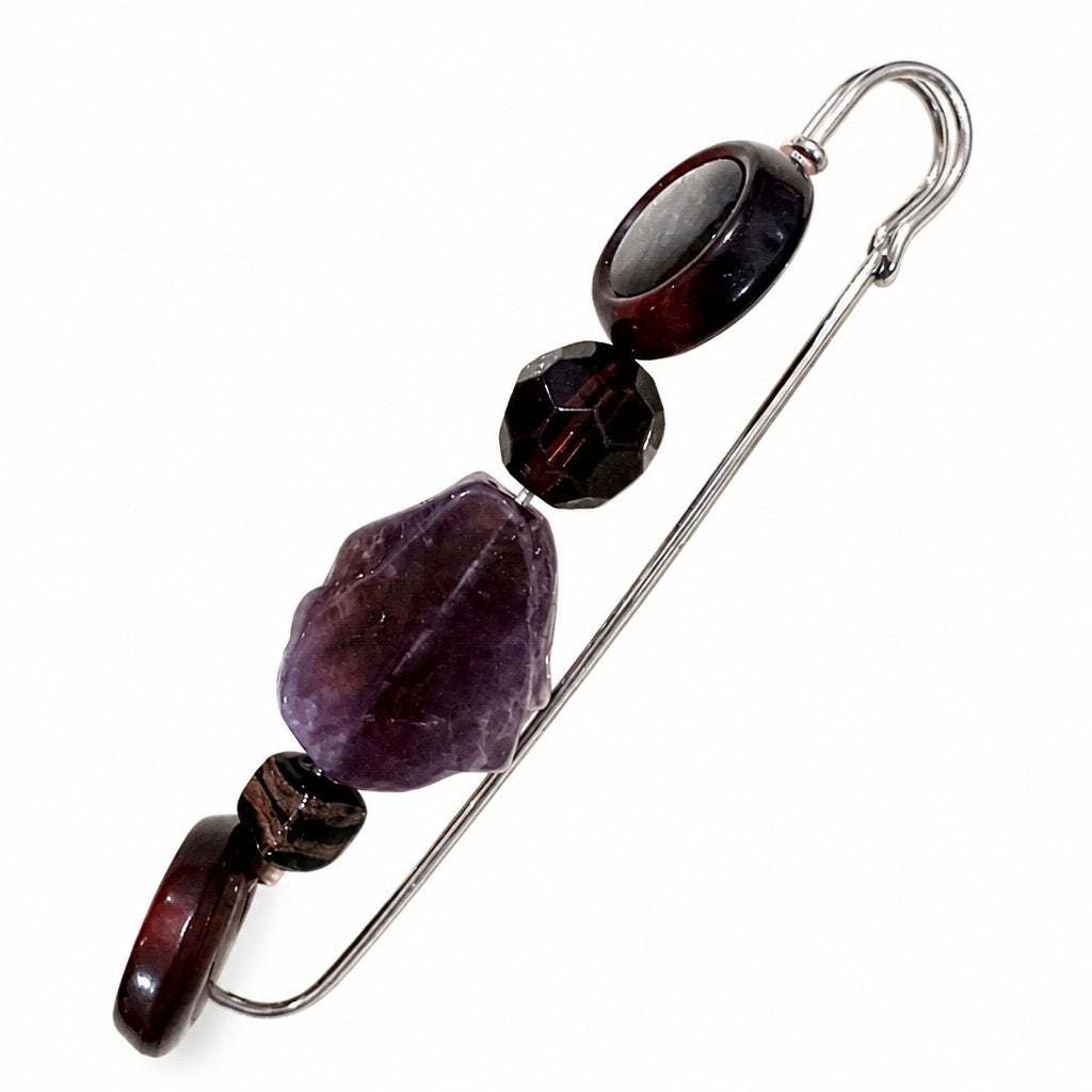 A silver-toned safety pin styled as a beaded brooch, featuring a large purple amethyst stone, a faceted dark red stone, and smaller dark brown and black beads, set against a plain white background. Silvia Tolomei best handmade jewelry in Tuscany. Tuscany jewelry. Tuscan jewelry. Populonia best store to visit. Things to do in populonia. 