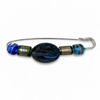 A handmade wire safety pin brooch decorated with glass beads, featuring a large central dark blue oval bead, a smaller blue bead on the left, a metallic olive-green cylinder, a textured bronze cylinder, and a small blue and white marbled bead on the right, set against a plain white background. Silvia Tolomei best handmade jewelry in Tuscany. Tuscany jewelry. Tuscan jewelry. Populonia best store to visit. Things to do in populonia. 