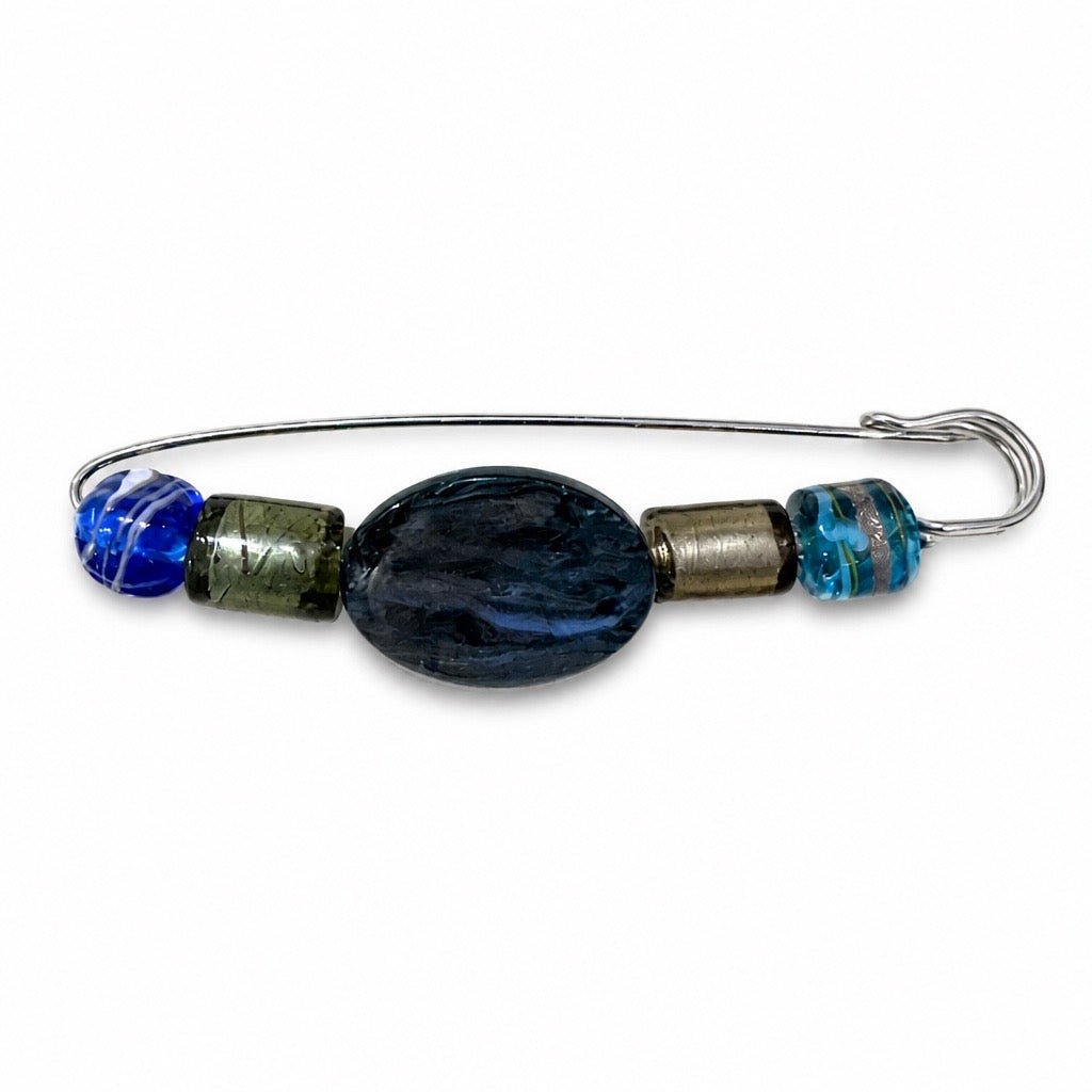 A handmade wire safety pin brooch decorated with glass beads, featuring a large central dark blue oval bead, a smaller blue bead on the left, a metallic olive-green cylinder, a textured bronze cylinder, and a small blue and white marbled bead on the right, set against a plain white background. Silvia Tolomei best handmade jewelry in Tuscany. Tuscany jewelry. Tuscan jewelry. Populonia best store to visit. Things to do in populonia. 