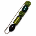 A handmade beaded pin featuring olive green textured beads, dark green cylindrical beads, a metallic gold-toned cylinder, and a black stone bead, all attached to a silver-tone metal safety pin. Silvia Tolomei best handmade jewelry in Tuscany. Tuscany jewelry. Tuscan jewelry. Populonia best store to visit. Things to do in populonia. 