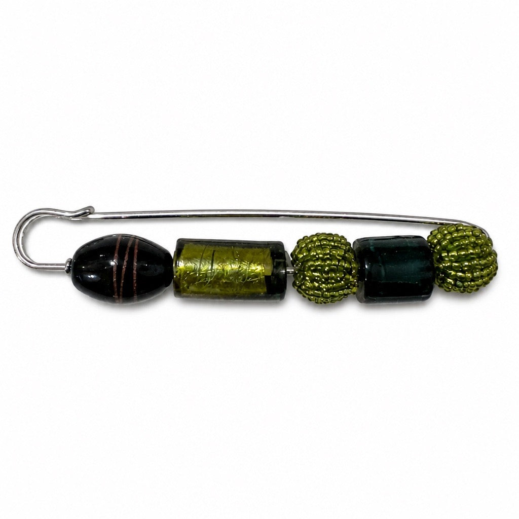 A safety pin brooch featuring decorative glass beads, including a black bead with gold stripes, a textured olive-green cylinder, two dark green beads, and two olive-green beaded spheres, on a white background. Silvia Tolomei best handmade jewelry in Tuscany. Tuscany jewelry. Tuscan jewelry. Populonia best store to visit. Things to do in populonia. 