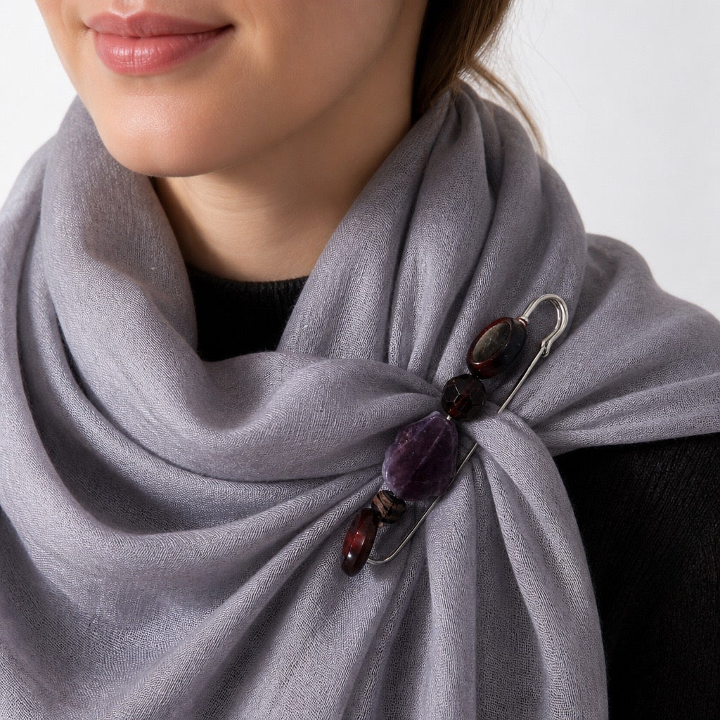 A close-up of a person wearing a soft gray wrap or scarf tied at the chest with a decorative brooch made of dark polished stones and a silver safety pin. Silvia Tolomei best handmade jewelry in Tuscany. Tuscany jewelry. Tuscan jewelry. Populonia best store to visit. Things to do in populonia. 
