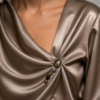 Close-up of a person wearing a satin-like draped top in a taupe-brown color, with a decorative brooch pinned at the front.