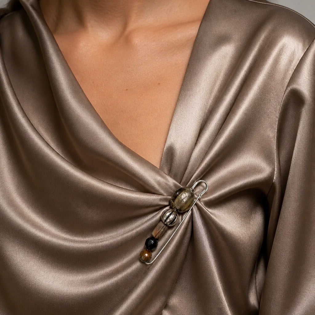 Close-up of a person wearing a satin-like draped top in a taupe-brown color, with a decorative brooch pinned at the front.