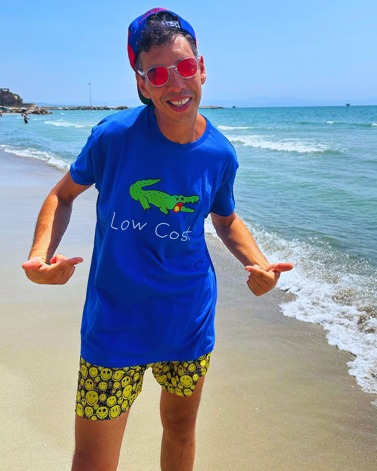 A man standing on a sandy beach wearing a bright blue t-shirt with a green alligator graphic and the text 'Low Cost', paired with yellow smiley-face patterned shorts, red sunglasses, and a blue cap.