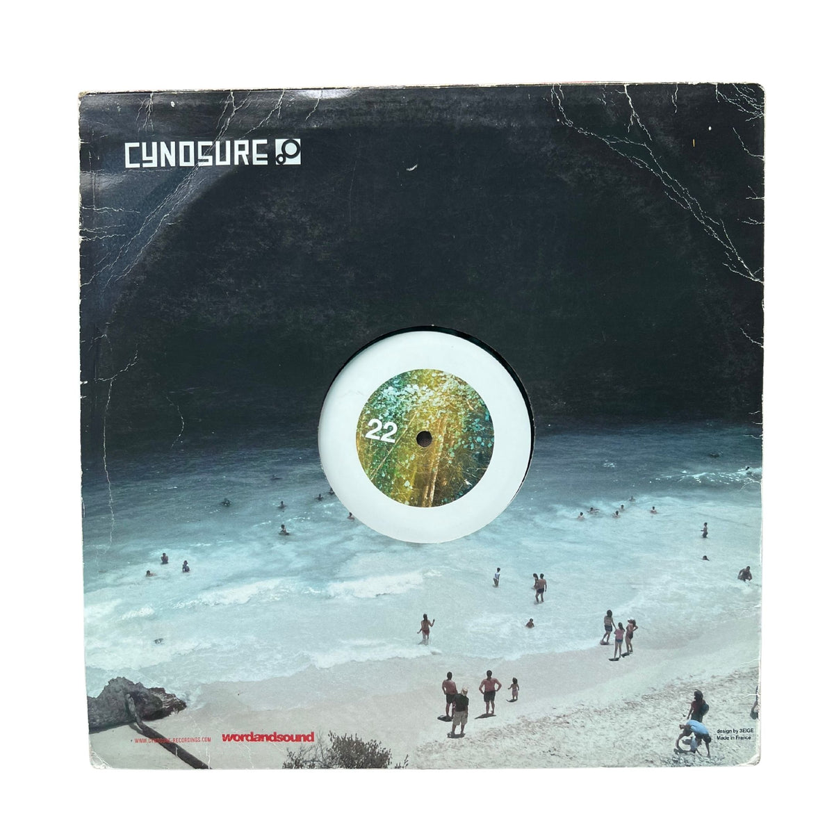 A square vinyl record sleeve for Cynosure's 'Wetlandsound' 22-inch record. The cover art features a surreal beach scene under a massive dark cave, with a white circular cutout in the center showing the green and yellow patterned center label of the vinyl record.