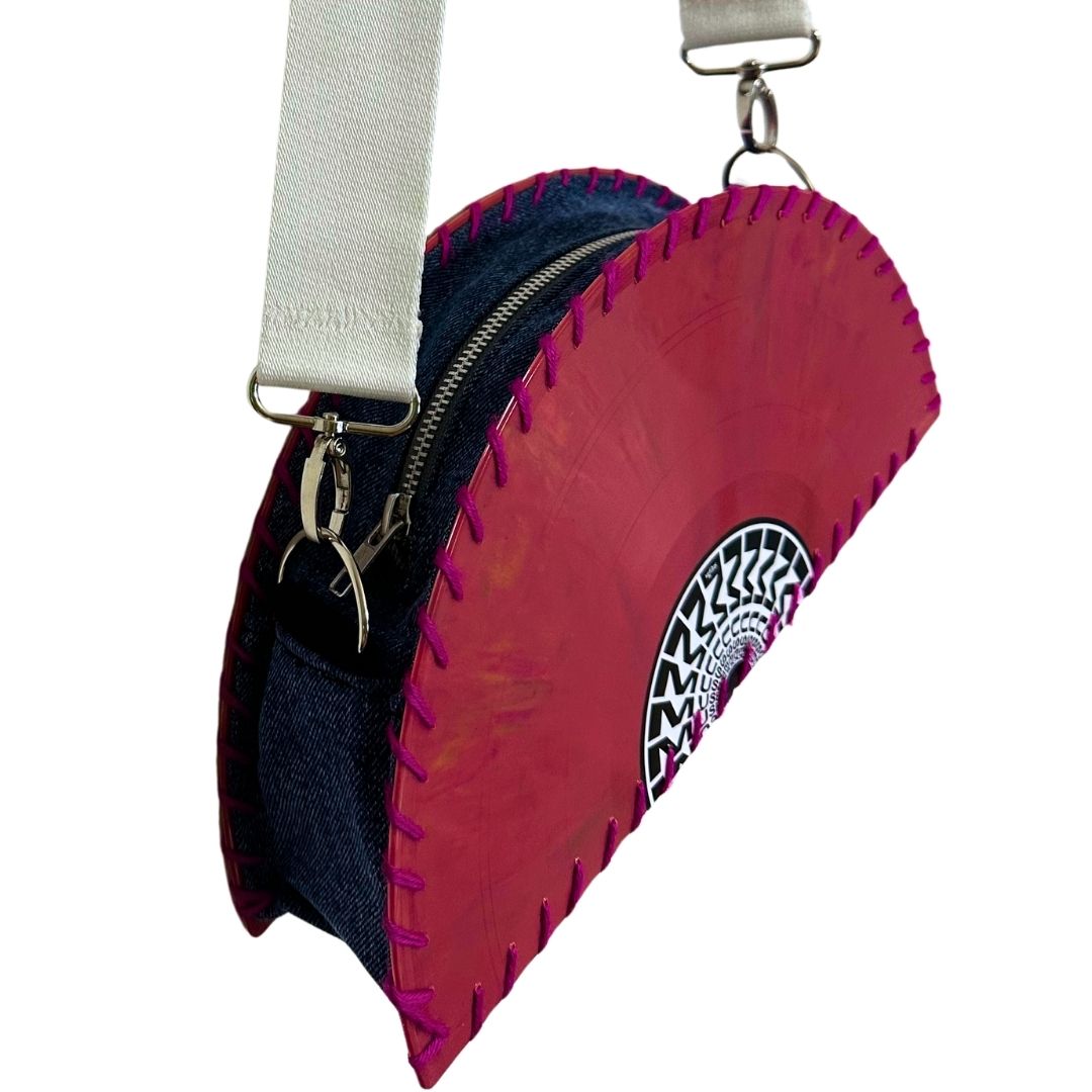 A small crossbody bag with a bright pink vinyl record design, dark navy blue denim side panels, magenta stitching, a white canvas strap, and a black-and-white circular graphic on the front, shown on a plain white background.