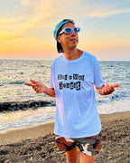 A man wearing a light blue graphic t-shirt with 'Non a Mai Graphik' printed on the front, standing on a pebble beach with the ocean and sunset in the background.