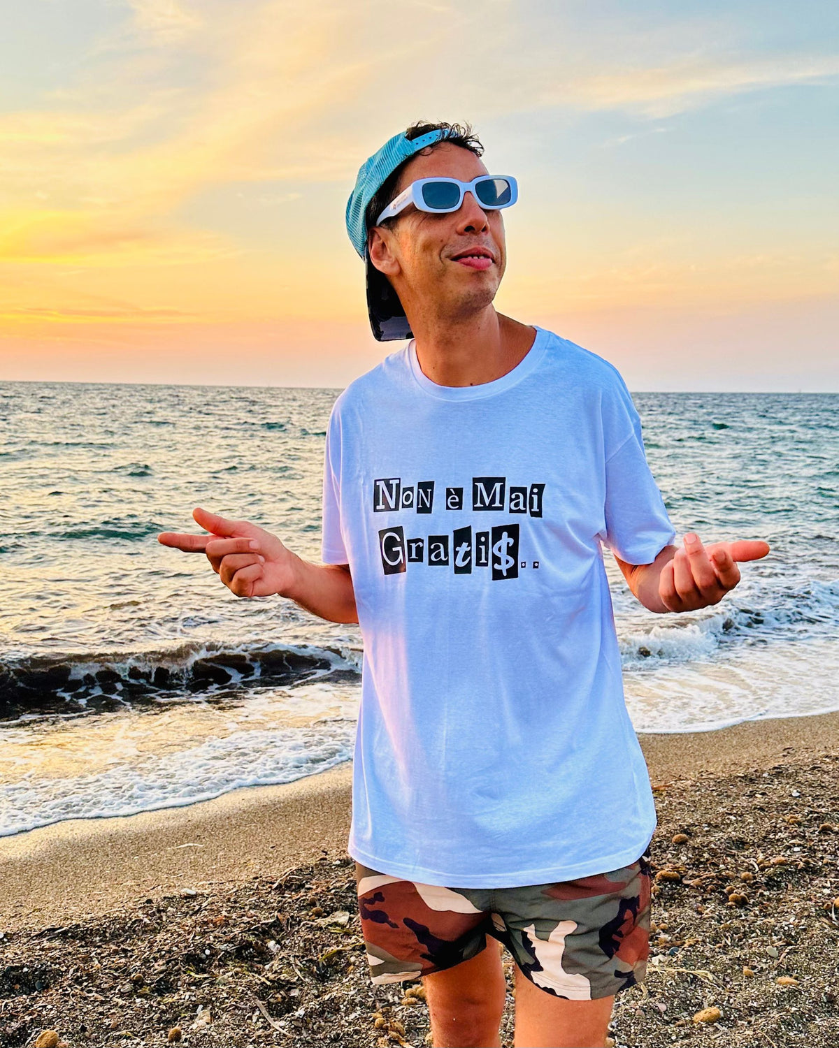 A man wearing a light blue graphic t-shirt with 'Non a Mai Graphik' printed on the front, standing on a pebble beach with the ocean and sunset in the background.