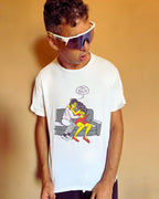 A man wearing a white graphic t-shirt with a cartoon illustration of a couple on a couch and a speech bubble that says 'OH SHIT.' He is also wearing white wrap-style sunglasses with a purple reflective lens, posed against a solid warm beige background.