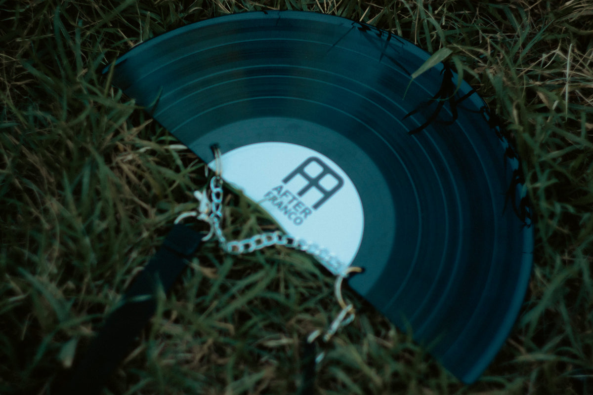 A black vinyl record-shaped hand fan with a silver chain, featuring the 'After Punk' logo, resting on green grass. AFTER FRANCO