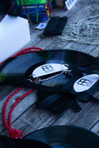A close-up of a black vinyl record necklace with a silver chain and a white circular label featuring the 'AA' logo of Aesthetic Apparel, resting on a weathered gray wooden surface. AFTER FRANCO