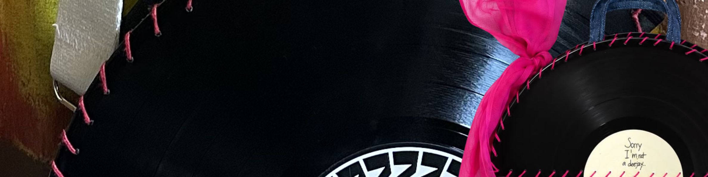 A close-up of a black sombrero-style hat with pink stitched trim and a bright pink band, partially obscuring a black vinyl record with a white label.