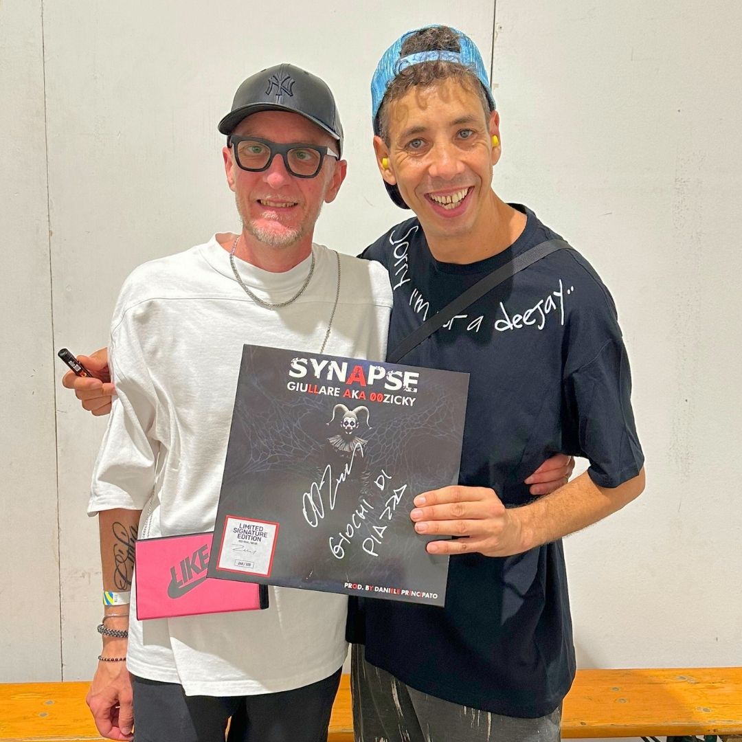 Two men holding a signed record with 'Synapse' on it, standing against a plain wall.SYNAPSE GIULLARE AKA 00ZICKY SORRY IM NOT A DEEJAY ANDREA DECUZZI.