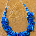 A vibrant blue beaded necklace made of various shell and button-like elements, displayed on a sandy beach background. Silvia Tolomei best handmade jewelry in Tuscany. Tuscany jewelry. Tuscan jewelry. Populonia best store to visit. Things to do in populonia. 