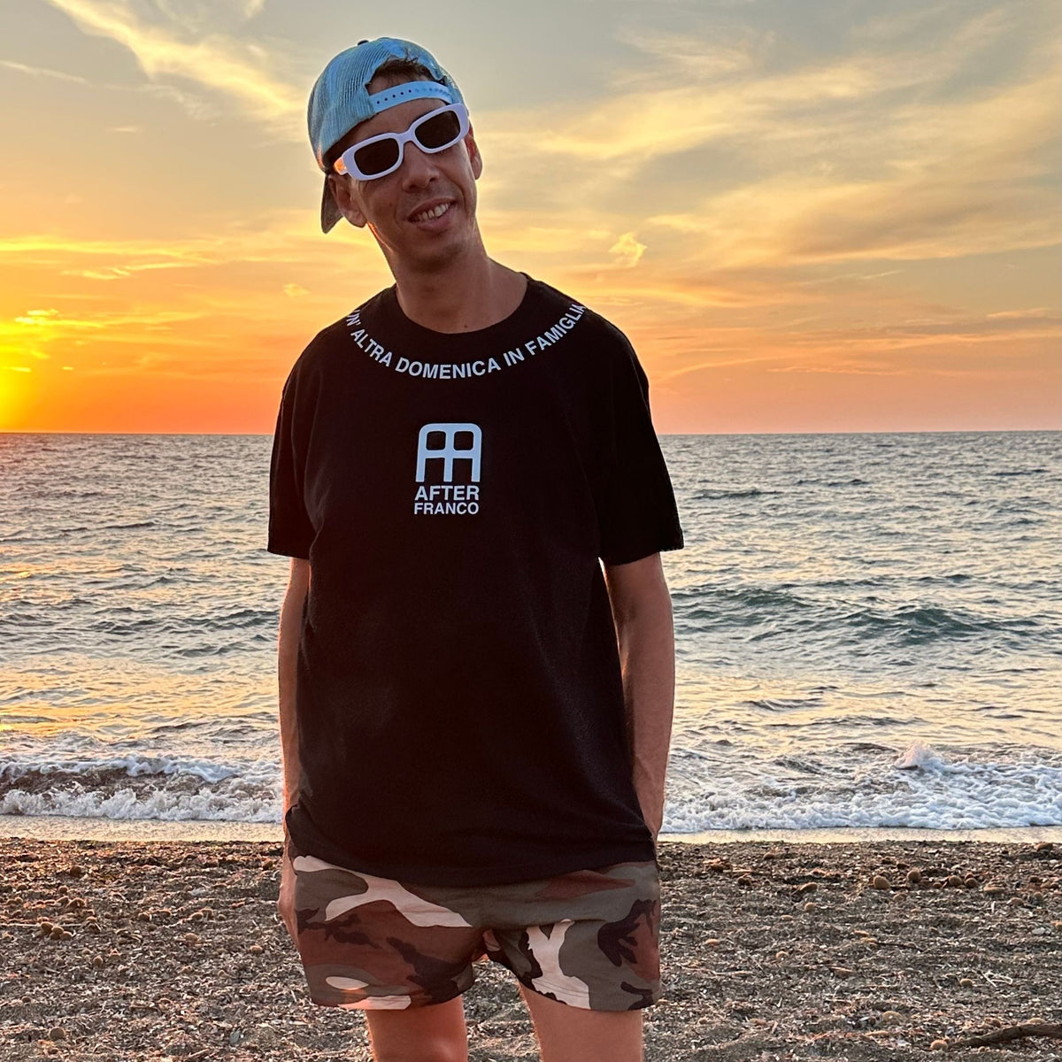A man standing on a beach at sunset wearing a black t-shirt with a white graphic and text, camouflage shorts, and a blue cap.