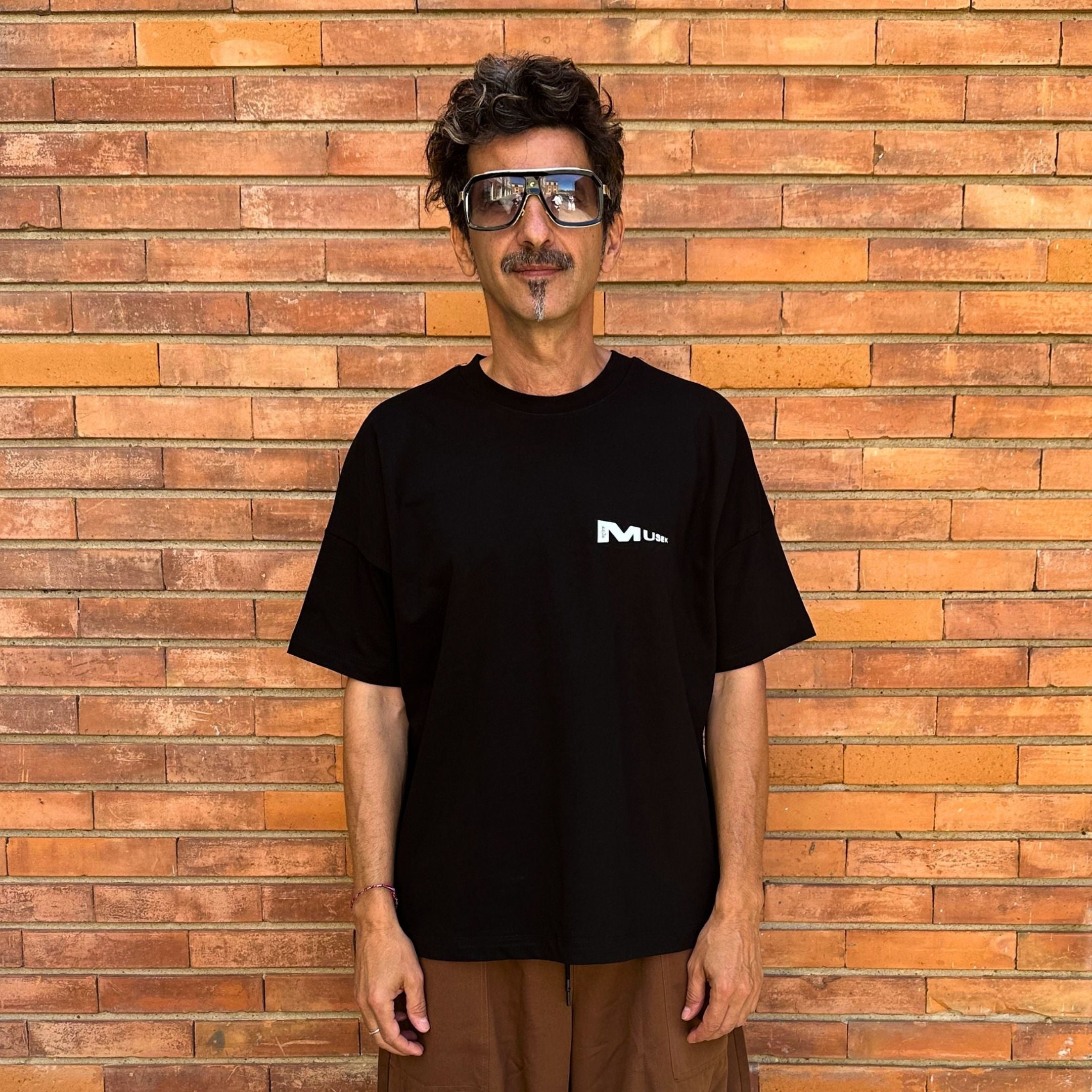 Man wearing a black t-shirt with a logo in front of a brick wall. Francesa Farfa black musek label t shirt