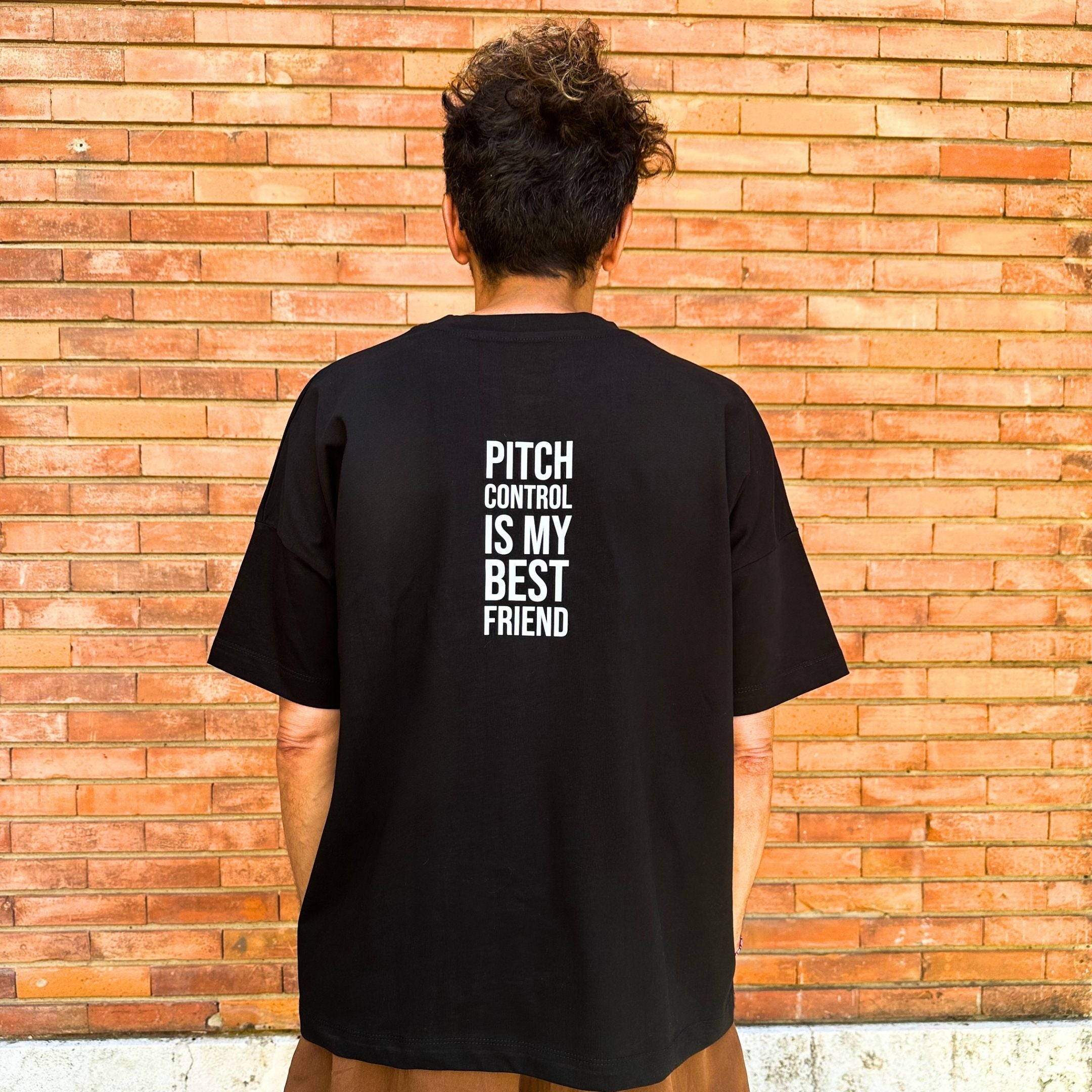 Person wearing a black t-shirt with 'Pitch Control is My Best Friend' text on the back, standing against a brick wall. Man wearing a black t-shirt with a logo in front of a brick wall. Francesa Farfa black musek label t shirt