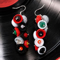 A pair of handmade dangle earrings featuring colorful felt-like rings and faux gemstones in red, black, white, green, and orange, displayed on a black vinyl record background. populonia